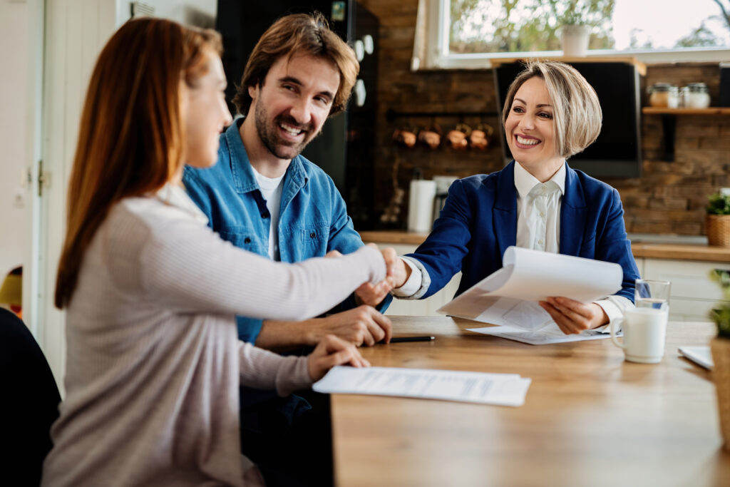 Happy real estate agent greeting young couple on a meeting at their home. Women are shaking hands.